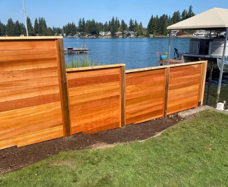 A wooden privacy fence stands beside a grassy lawn, overlooking a lake with docks and boats in the background.