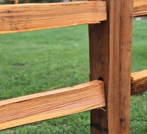 Close-up of a wooden fence post and rails with a grassy lawn and house in the blurred background.